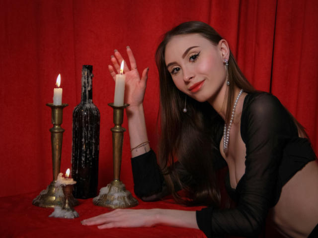 SaraQuincy with long brown hair, wearing a sheer black top, pearl necklace, and earrings, leans on red cloth, hand raised, next to lit candles and a black bottle.