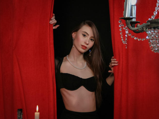 SaraQuincy, con lunghi capelli castani, rossetto rosso, reggiseno nero e una collana, che tiene delle tende rosse, orecchini d'argento, una candela a sinistra e un lampadario di cristallo a destra, in una stanza buia..