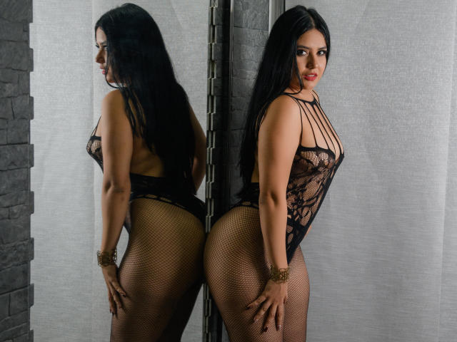 SarithaCandi with long black hair, wearing a black lace bodysuit and fishnet stockings, standing against a mirror, showing her big ass and small tits, with a confident smile.