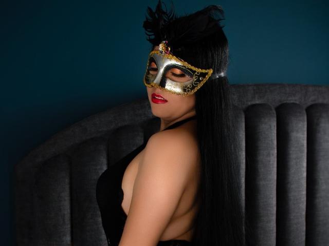 SarithaCandi with long black hair, wearing a gold and black mask with feathers, red lipstick, and a black dress with exposed back, sitting on a gray upholstered chair.