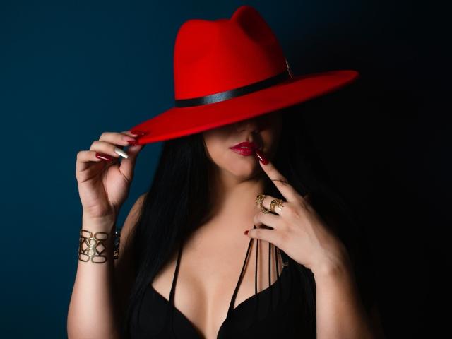 SarithaCandi mit schwarzen Haaren, rotem Hut, schwarzem Kleid, rotem Lippenstift, die mit dem Finger ihre Lippen berührt, einem goldenen Ring und Armband, wobei ihre Brüste sichtbar sind. Dunkler Hintergrund..