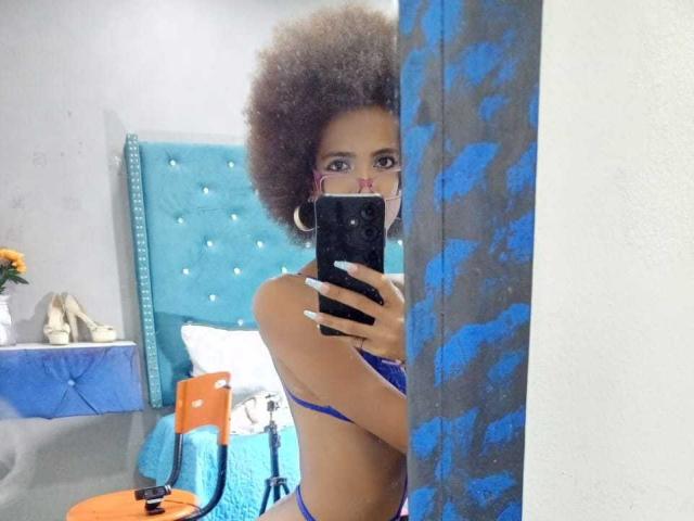 CanelaHott69 with big afro, wearing blue lingerie, holding phone in mirror selfie. Boobs visible, standing in front of blue-patterned wall, taking photo in bedroom.
