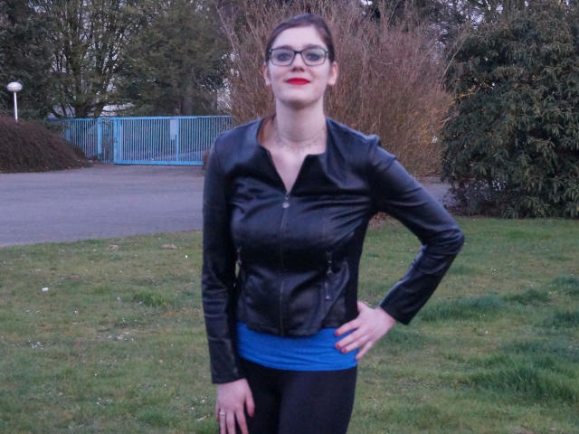 Zara-Bizarr, avec rouge à lèvres, veste en cuir noir, chemise bleue, pantalon noir, debout, les mains sur les hanches, lunettes, herbe verte, buisson et clôture bleue en arrière-plan..