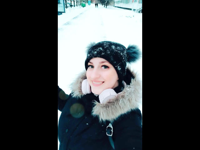 ChristineLove, com neve no gorro preto, vestindo um casaco preto com gola de pele e fones de ouvido brancos, sorrindo para a câmera em um ambiente urbano coberto de neve..