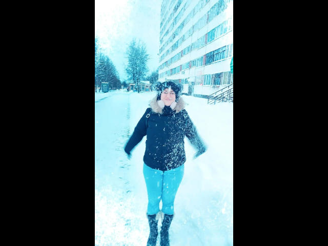 ChristineLove steht in einem verschneiten Stadtgebiet, trägt eine schwarze Jacke, blaue Hosen und schwarze Stiefel, hat Schneeflocken im Gesicht und auf den Armen und blickt nach vorne..