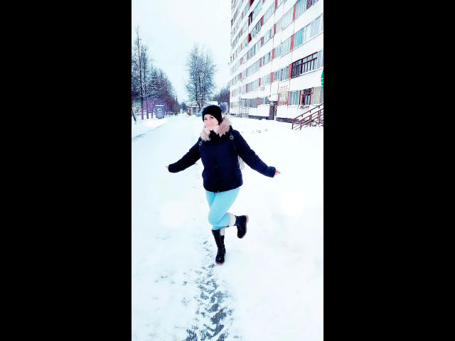 ChristineLove-hot rent door de sneeuw, gekleed in een zwarte jas, een lichtblauwe rok, zwarte laarzen en een zwarte hoed. Haar armen zijn uitgestrekt en ze heeft een vrolijke uitdrukking..