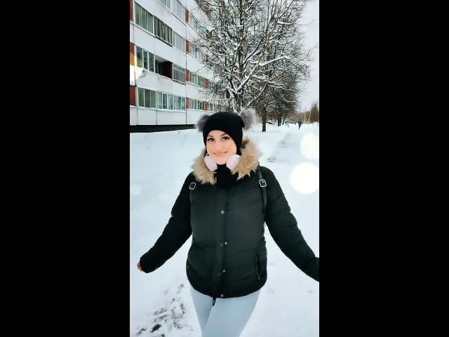 ChristineLove, belle et souriante, portant une veste noire matelassée avec un col en fourrure, un chapeau noir avec des pompons blancs, des protège-oreilles roses, et se tenant dans une zone urbaine enneigée..