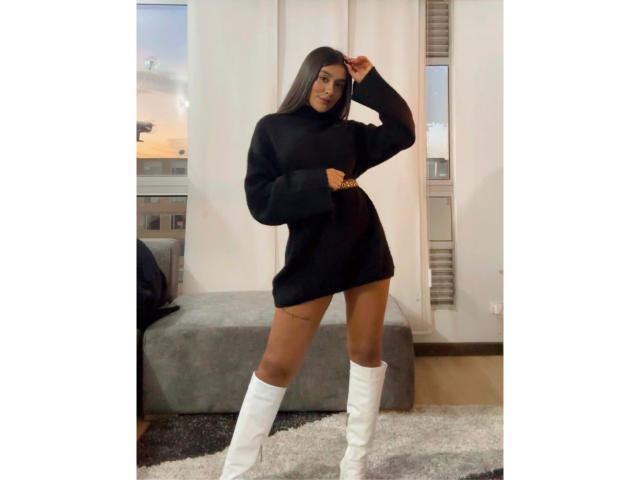 ThaliaAres, with long dark hair, wearing a black turtleneck sweater dress, white knee-high boots, standing in a room, hand on head, showing a tattoo on her right thigh.