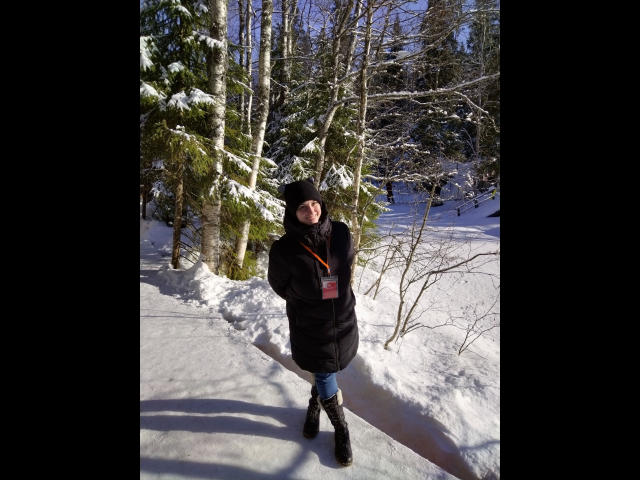 ChristineLove-hot se tient dans une forêt enneigée, vêtue d'un manteau noir matelassé, d'un chapeau noir et de bottes noires, avec une sangle rouge visible, et sourit à l'appareil photo..