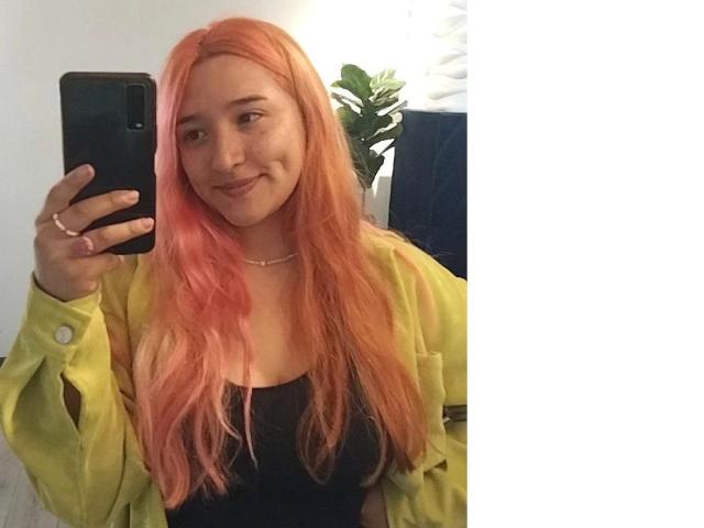 IsabellaClown with pink hair, smiling, holding a phone, wearing a yellow-green jacket, black top, and pink ring, taking a selfie, showing her tits subtly with her jacket.