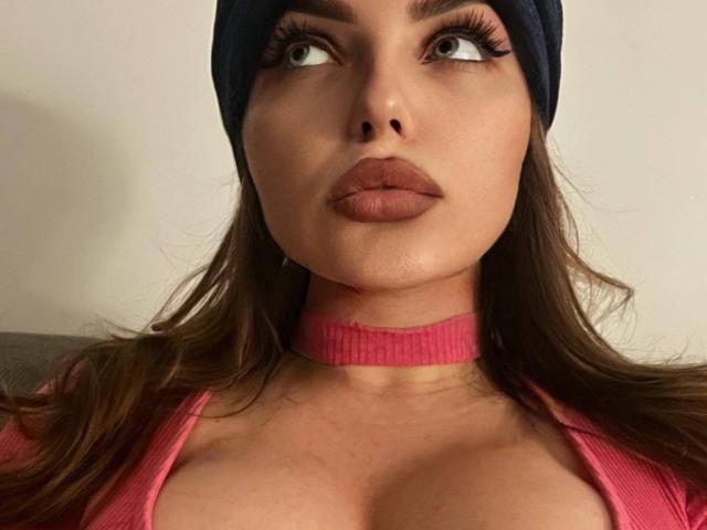 JossephinRose tem olhos verdes grandes, lábios rosados cheios e cabelo castanho ondulado. Ela está usando um colar rosa e um gorro azul, com seus seios visíveis em uma blusa rosa..