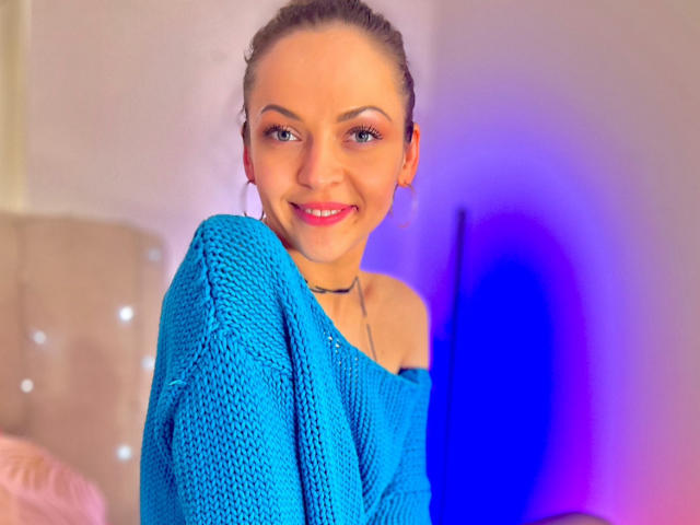 Veronika Dylan, com cabelo castanho claro, olhos azuis e batom vermelho, vestindo um suéter azul de tricô com um ombro descoberto, sorrindo para a câmera. Seus seios estão parcialmente cobertos..