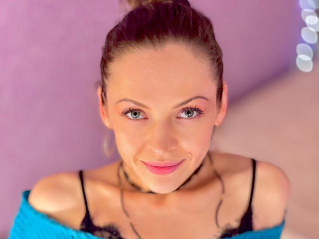 VeronikaDylan, aux yeux bleus, avec les cheveux bruns attachés en chignon, portant un soutien-gorge en dentelle noire et un collier, souriant à la caméra, nue, montrant ses seins et un petit collier..
