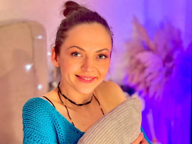 Veronika Dylan, com o cabelo escuro preso em um coque, sorrindo, vestindo uma blusa azul de tricô, segurando um travesseiro cinza, usando um colar preto, e com iluminação rosada-azul no fundo..