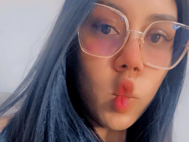 StephanieDavies with blue hair, pouty lips, and transparent glasses, looking directly at the camera, has a slightly flushed face, and is wearing a simple top.
