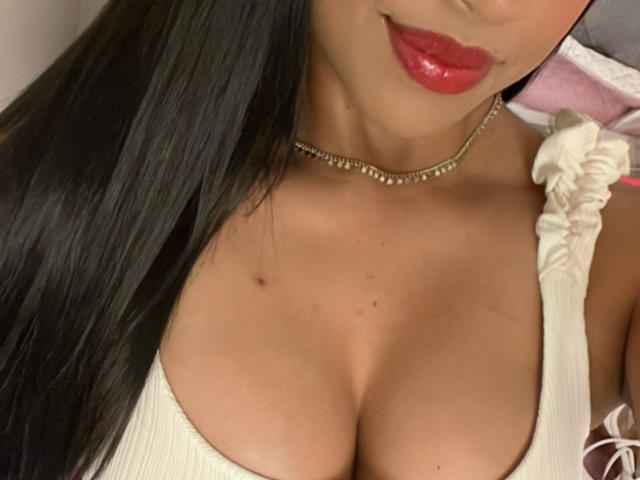 Stephanie Davies, com lábios vermelhos, cabelos pretos longos, colar de ouro e uma blusa branca de decote baixo que mostra seus seios grandes e redondos e uma pequena pinta no peito..