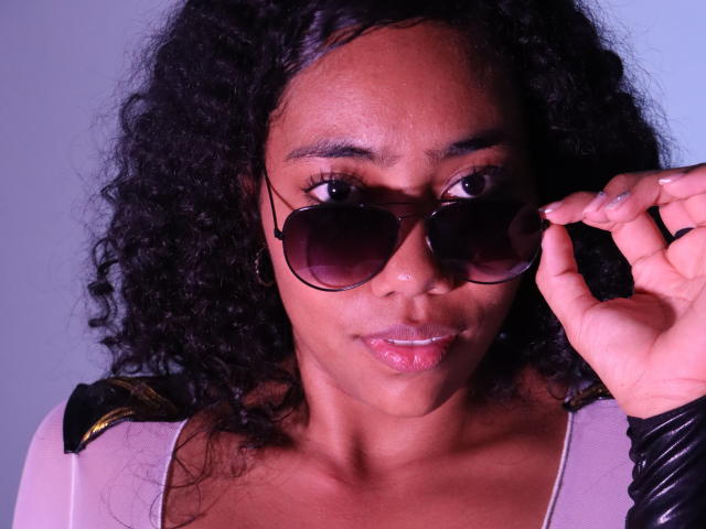 AishaRhodes with curly black hair, brown skin, wearing dark sunglasses, holding them with her right hand, pinkish-lip gloss, and a beige top with black shoulder accents.