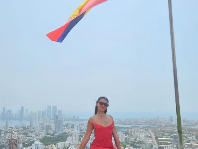 AlessiaFerry with black hair, sunglasses, and a red off-shoulder dress, standing in front of a flying Colombian flag, showing her tits and ass. Cityscape in the background.