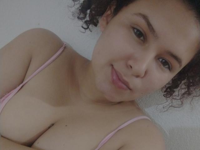 PaoleHot with curly brown hair, light skin, and pink bra, showing her big tits and subtle smile. She's looking at the camera with a gentle expression.