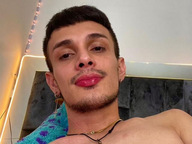 SlimLewis with short black hair, light brown skin, small goatee, red lips, black ear studs, gold necklace, and a shirtless upper body, looking into the camera.