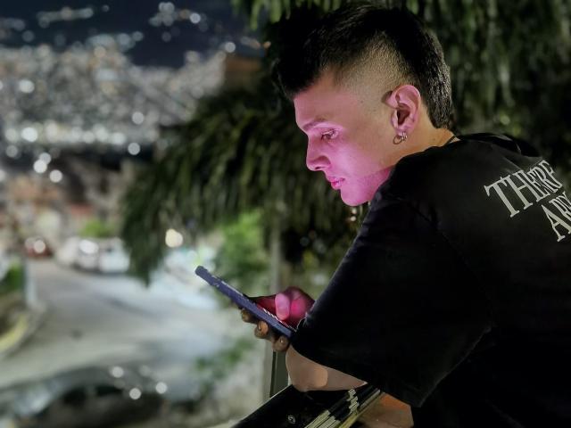 SlimLewis with a mohawk, black shirt, ear piercing, and pink lighting, leaning on a wall, staring at a phone, holding a drink, urban night setting.
