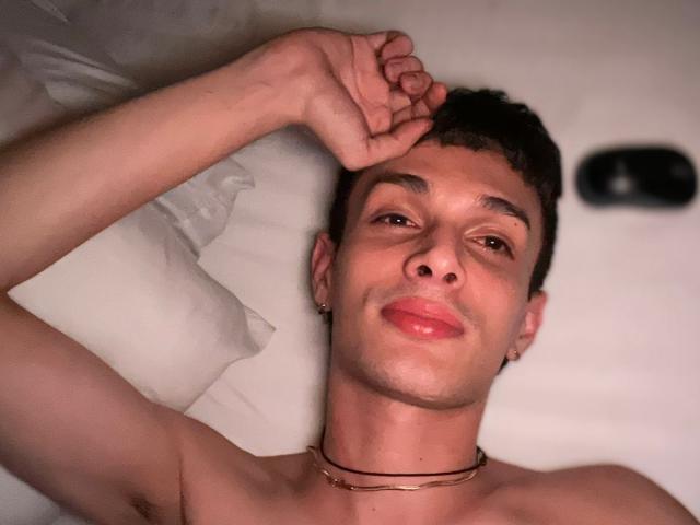 SlimLewis with short brown hair, ear piercings, and a gold choker, lying on a bed, arm above head, smiling with red lips, looking relaxed. Soft light on his face.
