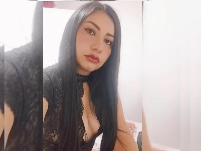 Una joven con cabello negro largo, vestida con una blusa negra de encaje que deja ver parte del escote, lleva lápiz labial rojo y está mirando en un espejo. Su reflejo se ve a la izquierda..