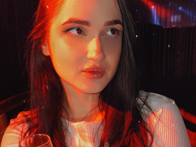 KerryB has shiny red-lit skin, long dark hair, and full pouty lips. She's wearing a white ribbed shirt, holding a glass, with a sultry, half-lidded gaze.