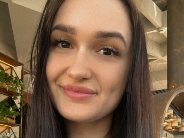Het gezicht van KerryB, met bruine ogen, lang donkerbruin haar, dikke wenkbrauwen en roze lippen, met een lichte glimlach, en een natuurlijke huid, op een selfie. Haar borsten zijn niet zichtbaar..