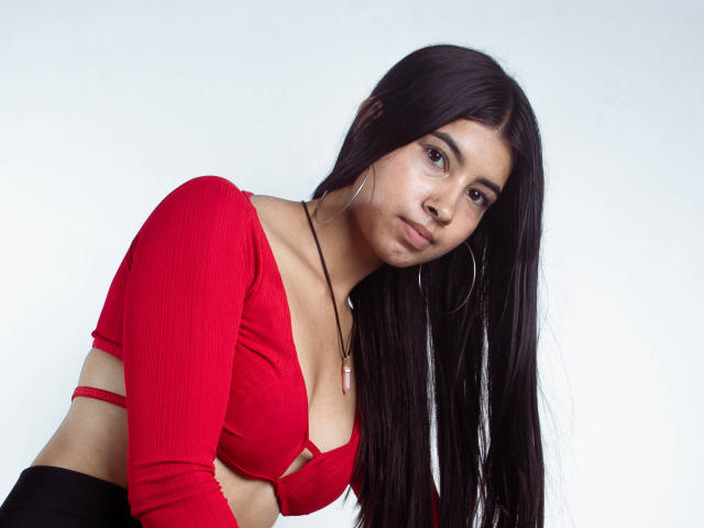 KarinaVughet, avec de longs cheveux noirs, un haut court rouge à rayures, de petites seins, un bas noir, des boucles d'oreilles cerceaux, un collier, penchée en avant, expression sérieuse, peau claire, fond blanc..