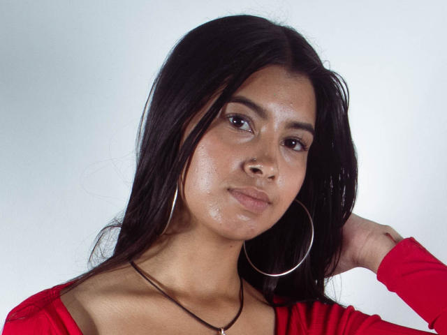 Karina, mit glatter Haut, dunklem, langem Haar, einem kleinen Nasenring, großen Reifenohrringen, einem roten Oberteil und einer Halskette, blickt leicht zur Seite, eine Hand im Haar..