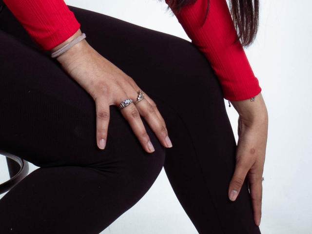 KarinaVughet met haar handen op zwarte broek, een rood shirt met lange mouwen aan, zilveren ringen zichtbaar aan haar vingers, zittend met gespreide benen, witte achtergrond..