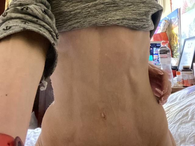 Kabblasha's slanke, getoonde middel met zichtbare ribben en een kleine navel, draagt een grijze shirt die omhoog is getrokken, waardoor een plat buikje zichtbaar is, en houdt in de achtergrond een fles vast..