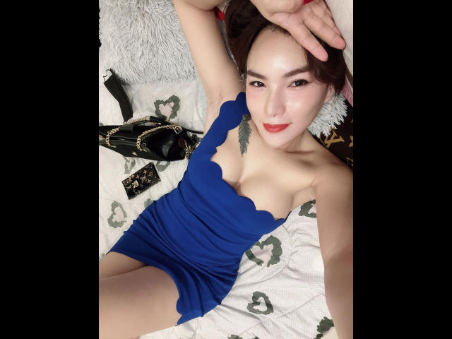 NaughtyAzumi69 - sensuale in un abito blu senza spalline, sdraiata su un letto, con le braccia alzate, con una borsa nera e una tracolla accanto a lei, sorridente, che mostra piccoli seni e un viso carino..