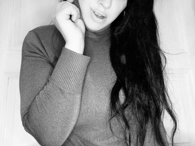 Raubmiez-hot mit langem, dunklem Haar, trägt einen grauen, gerippten Pullover, hält ihr Kinn mit einer Hand, lächelt leicht, Schwarz-Weiß-Bild..