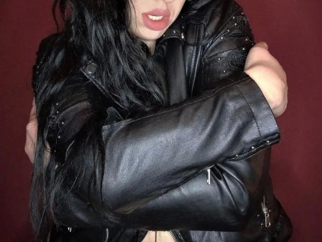 Raubmiez-hot, con chaqueta de cuero negro, brazos cruzados, cabello negro largo, labios rosados ligeramente entreabiertos, dientes visibles, fondo rojo. Los senos y los brazos están cubiertos por la chaqueta..