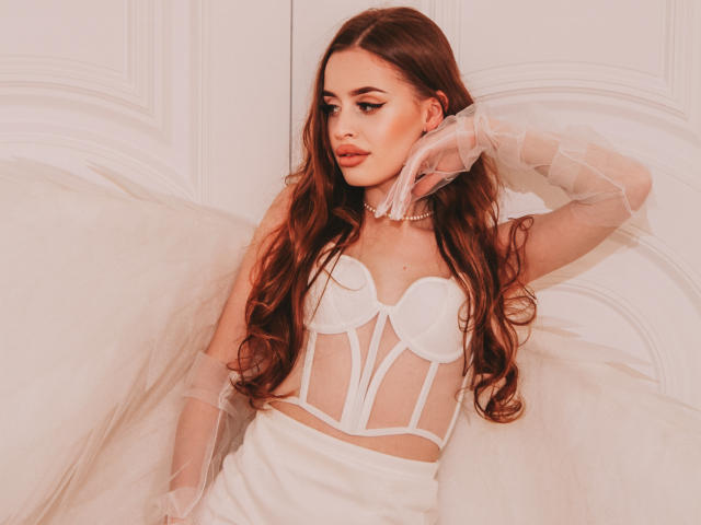 CherylFoxy with long brown hair, wearing a sheer white bustier that exposes her small tits, and a fluffy white skirt, with a transparent glove and pearl necklace.