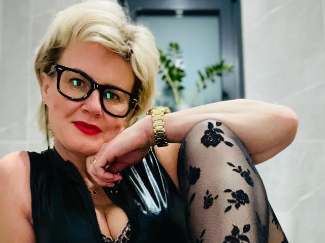 AdeleMILF-hot with short blonde hair, black glasses, red lipstick, black floral tights, gold watch, and black top, leaning on her knee, showing her tits.