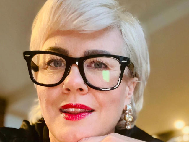 AdeleMILF-hot with short blonde hair, black glasses, red lipstick, and pearl earrings, smiling with a hand under her chin, showing a green reflection in her glasses.