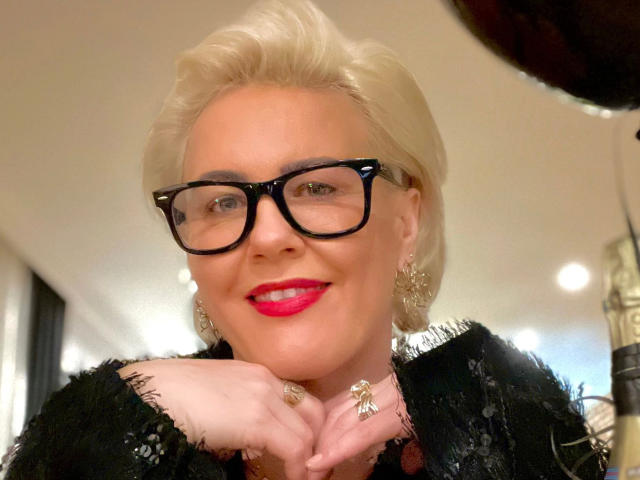 AdeleMILF-hot with short blonde hair, black glasses, red lipstick, black fringed top, and earrings, smiling, hands under chin, looking confident. She's got a cute ass too.
