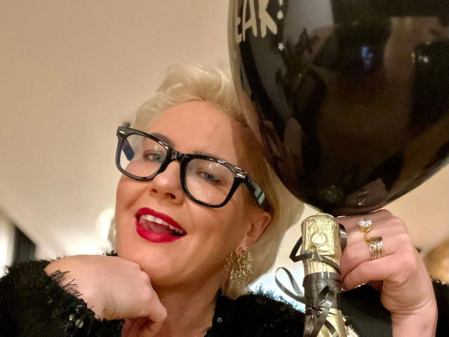 AdeleMILF-hot with blonde hair, black glasses, red lipstick, holding a gold wine bottle with a black glass, smiling. She's wearing a black lace top and earrings.