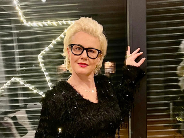 AdeleMILF-hot with blonde hair, black glasses, red lipstick, and black sequined fur top, waving. She's got big tits and a cute ass, standing in front of a glass door.