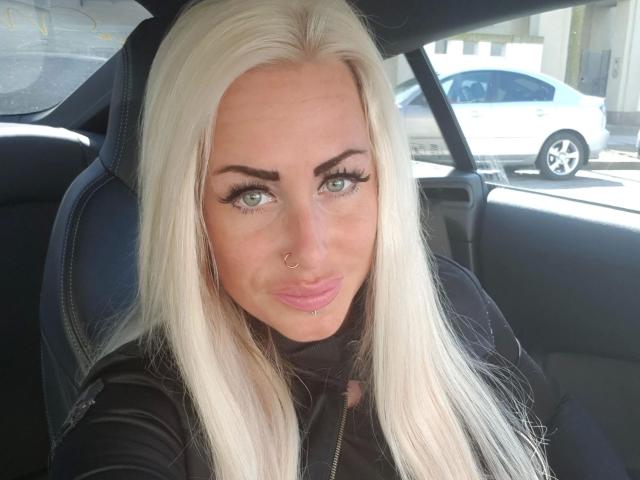 XXMissesPORXX-hot with platinum blonde hair, green eyes, nose ring, and black eyeliner, sitting in a car, wearing a black jacket, looking at the camera with a slight smile.