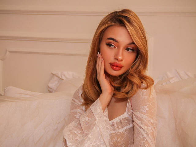 KylieBeaty with blonde hair, wearing a white lace top, hands on her face, looking seductive with red lips, sitting on a bed with white pillows.