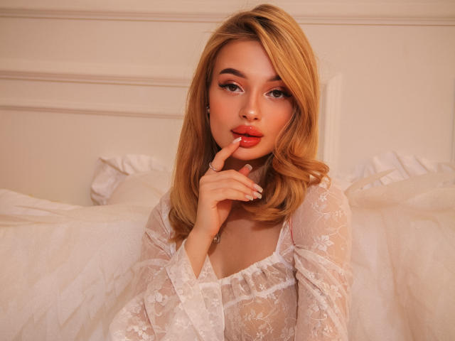 KylieBeaty with blonde hair, wearing a sheer white lace dress, has her finger slightly in her red lips, looking seductive with a cute, sexy, and attractive face.