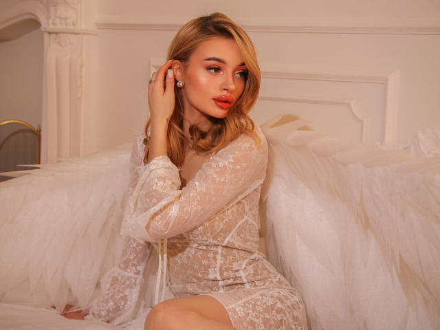 KylieBeaty with blonde hair, red lipstick, wearing a sheer lace dress, white wings, sitting on a bed, touching her ear, looking to the side, soft light.
