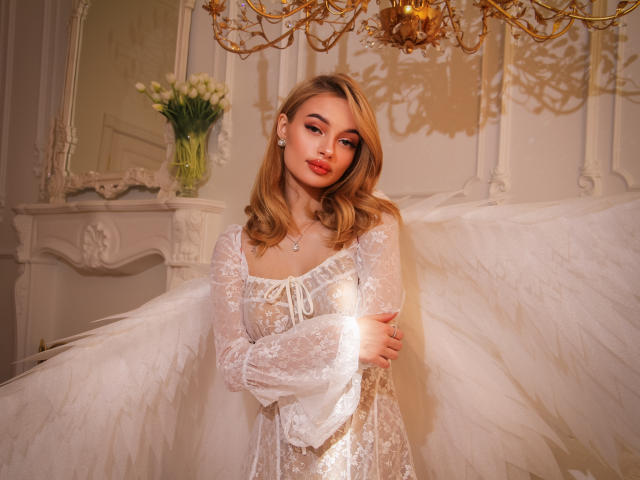 KylieBeaty with angel wings, wearing a white lace dress, standing in a room with a golden chandelier, holding her tits, looking cute with blonde hair and pink lipstick.