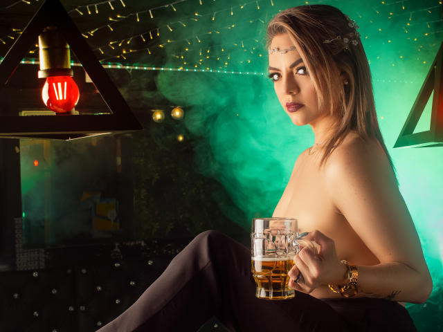 AvaMia, topless with medium tits, holding a glass of whiskey, wearing black pants and a gold bracelet, in a green-lit room, looking at the camera.