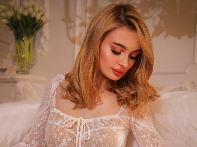 KylieBeaty with blonde hair, wearing a sheer white lace top, showing her tits, looking down with soft makeup and red lipstick, in a warm, cozy setting.