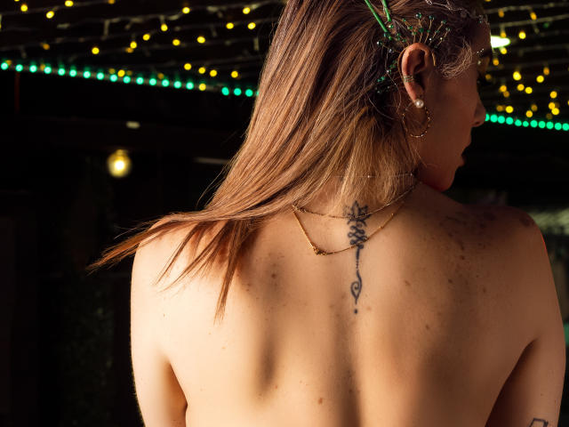 AvaMia's topless back, brown hair, green hairpins, small earrings, tattoo down her spine, light skin, slight freckles, dimly lit with yellow and green string lights.